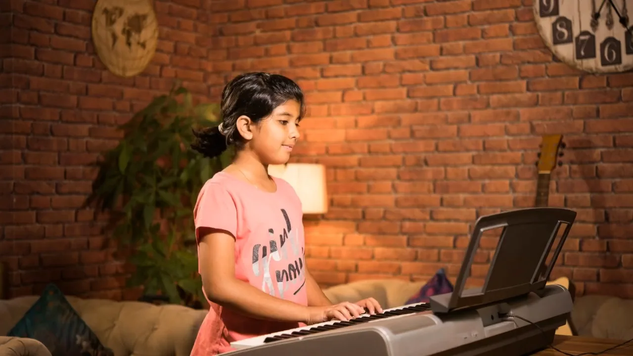 A young learner playing a keyboard in a warm, lamp-lit room during her online piano class